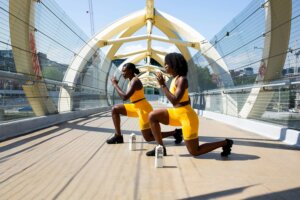Two women in yellow workout outfits doing lunges on a pedestrian bridge with Boxed Water cartons nearby