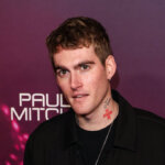 Man with tattoos wearing black jacket and sneakers posing on pink carpet at Paul Mitchell event backdrop