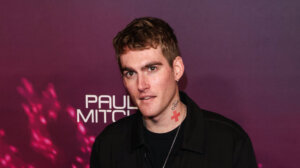 Man with tattoos wearing black jacket and sneakers posing on pink carpet at Paul Mitchell event backdrop