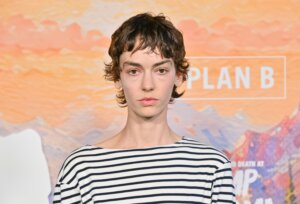 Person with short curly hair wearing a black and white striped shirt in front of a "PLAN B" backdrop