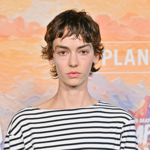 Person with short curly hair wearing a black and white striped shirt in front of a "PLAN B" backdrop