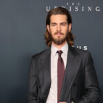 Andrew Garfield in a dark pinstripe suit with a maroon tie at The Uprising event backdrop