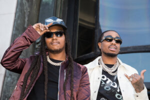 Rapper Takeoff wearing a purple jacket and cap next to rapper Quavo in a white jacket and sunglasses outdoors