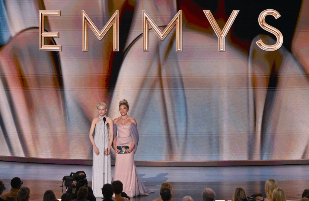 Two women in elegant gowns presenting onstage at the Emmy Awards with a large "EMMYS" sign behind them.