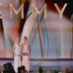 Two women in elegant gowns presenting onstage at the Emmy Awards with a large "EMMYS" sign behind them.