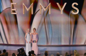 Two women in elegant gowns presenting onstage at the Emmy Awards with a large "EMMYS" sign behind them.