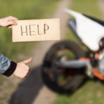 Person holding a cardboard sign reading "HELP" and giving a thumbs up near a dirt bike on a rural path.