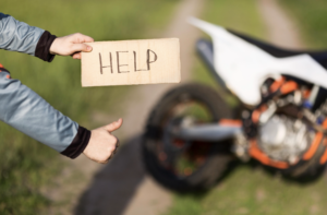 Person holding a cardboard sign reading "HELP" and giving a thumbs up near a dirt bike on a rural path.