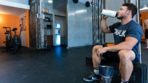 Man in Chalk t-shirt drinking from a shaker bottle next to a bag of Clean & Lean Recovery Shake in a gym.